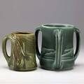 Rookwood two zline mugs one a loving cup with abstract design and emerald green glaze 1903 the other embossed with cornstalks and covered in brown and green glaze 1906 both stamped with flame ma